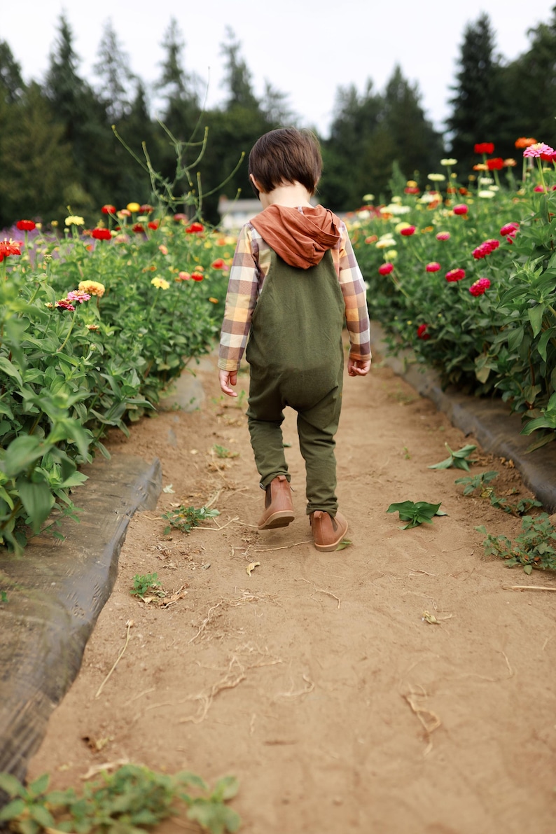 May include: A child wearing olive green overalls over a long-sleeved plaid shirt with a rust-colored hood, paired with brown leather boots. The child is walking away from the camera on a dirt path between rows of colorful zinnia flowers.