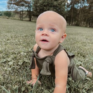 May include: A baby with bright blue eyes wearing an olive green romper featuring shoulder straps with tied knot details. The garment is a solid, muted green color with a simple neckline and sleeveless design, worn by a young child in a field.