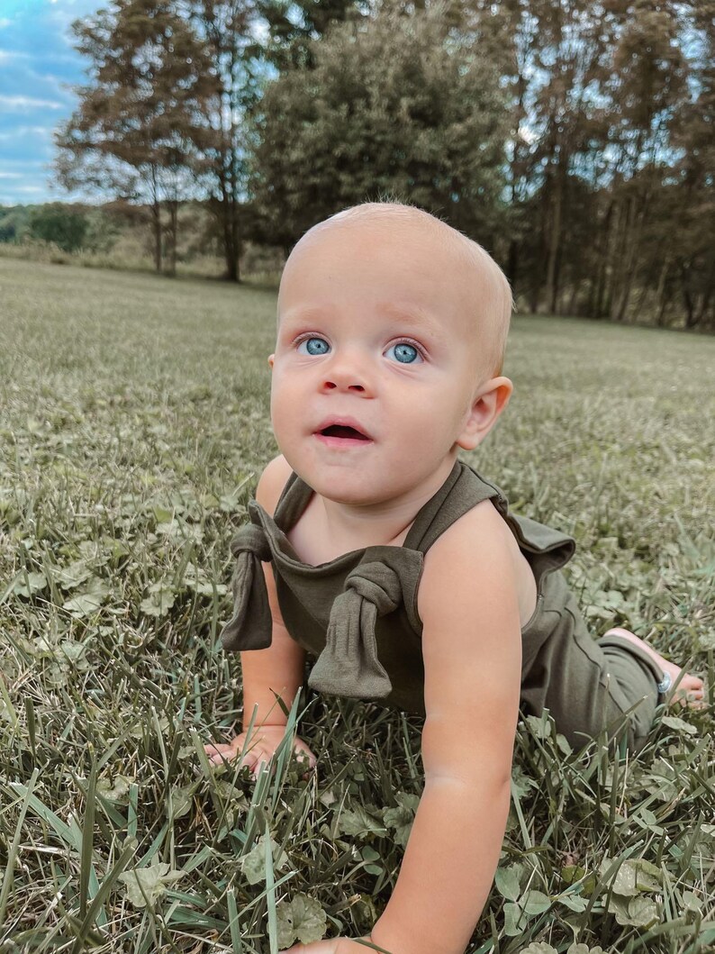 May include: A baby with bright blue eyes wearing an olive green romper featuring shoulder straps with tied knot details. The garment is a solid, muted green color with a simple neckline and sleeveless design, worn by a young child in a field.