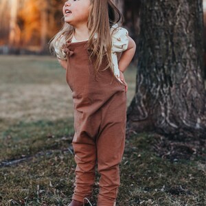 May include: A solid brown cotton overall with knotted shoulder straps worn over a cream-colored short-sleeved shirt featuring a small brown pattern. The outfit is completed with brown leather boots and a small matching hair clip in the hair.