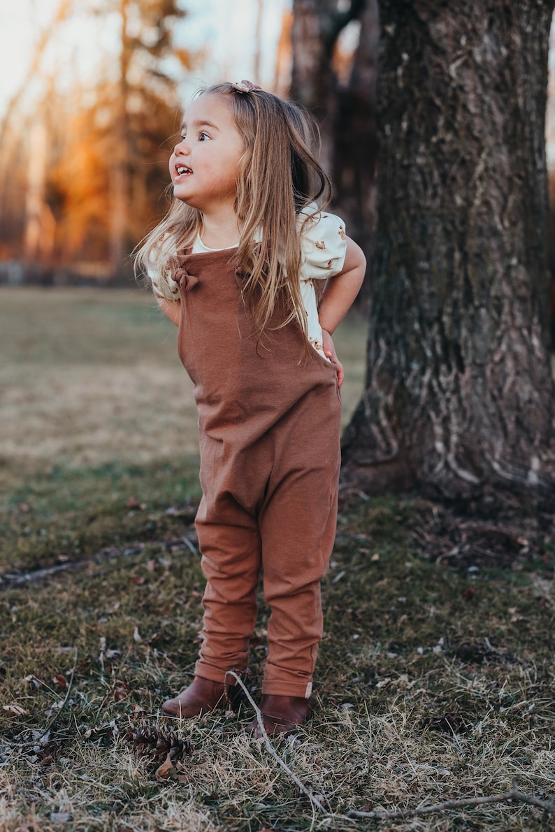 May include: A solid brown cotton overall with knotted shoulder straps worn over a cream-colored short-sleeved shirt featuring a small brown pattern. The outfit is completed with brown leather boots and a small matching hair clip in the hair.
