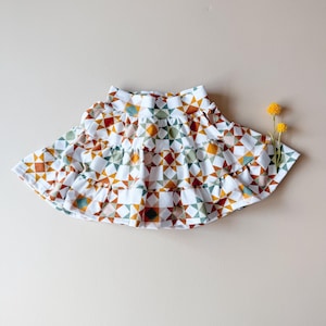 May include: A white tiered skirt with a geometric pattern in shades of orange, green, and brown. The skirt has a ruffled hem and is displayed on a neutral background. Two small yellow flowers are placed to the right of the skirt.