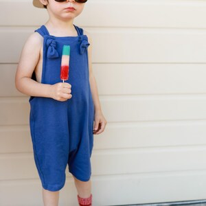 May include: A blue sleeveless romper with knotted shoulder straps, a beige baseball cap, dark-rimmed sunglasses, red and white patterned socks, and white sneakers. A tri-colored popsicle in green, white, and red is held in the hand.