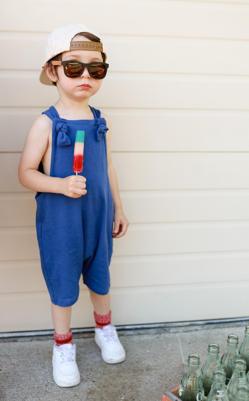May include: A blue sleeveless romper with knotted shoulder straps, a beige baseball cap, dark-rimmed sunglasses, red and white patterned socks, and white sneakers. A tri-colored popsicle in green, white, and red is held in the hand.