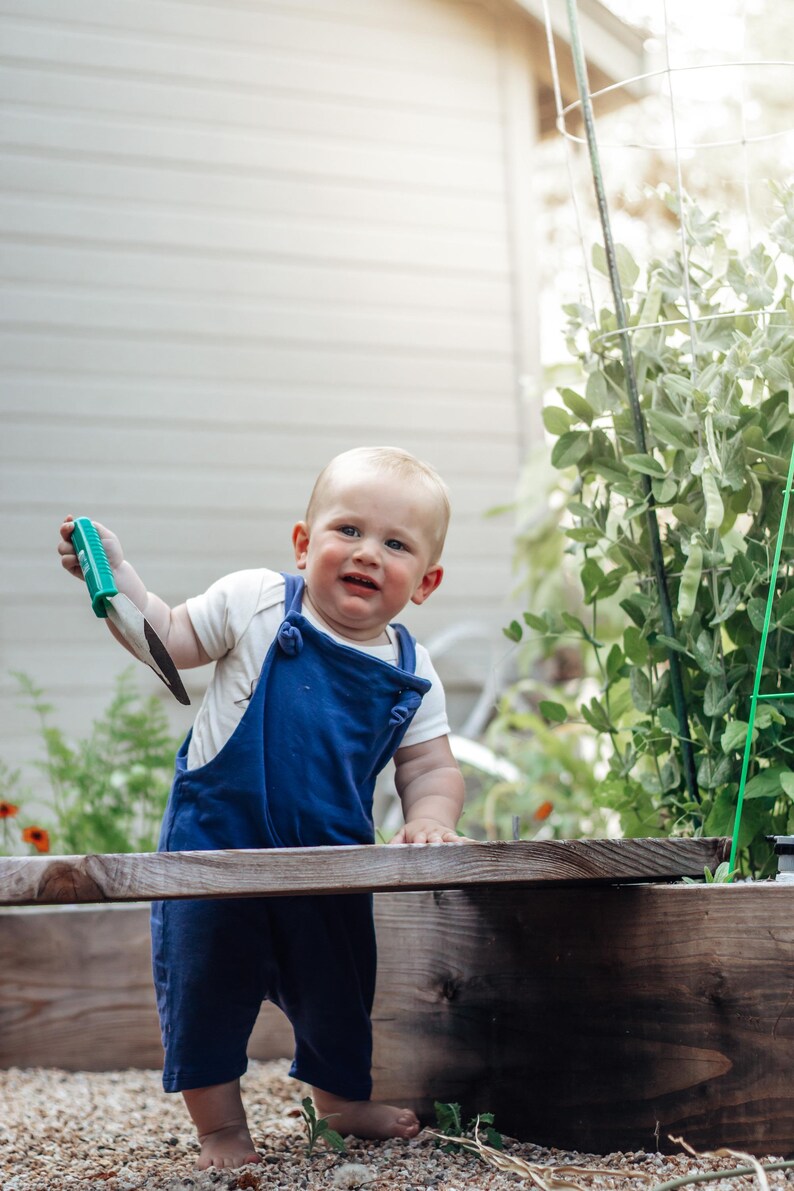 May include: A toddler wearing a white short-sleeved bodysuit and navy blue overalls, holding a small green-handled garden trowel with a metal blade. The child stands barefoot on gravel next to a wooden garden bed with green plants.