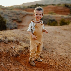 May include: A toddler wearing a tan-colored pair of overalls with shoulder straps and a short-sleeved white t-shirt featuring thin horizontal brown stripes and a solid brown crew neckline, paired with light brown leather shoes.