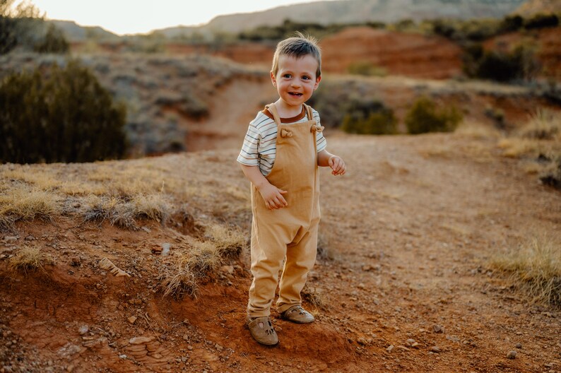 May include: A toddler wearing a tan-colored pair of overalls with shoulder straps and a short-sleeved white t-shirt featuring thin horizontal brown stripes and a solid brown crew neckline, paired with light brown leather shoes.