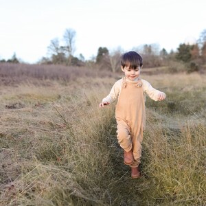 May include: A toddler wearing a light beige long-sleeved shirt underneath tan overalls with knotted shoulder straps and brown leather boots. The clothing is solid-colored and features a simple, functional design suitable for outdoor movement.