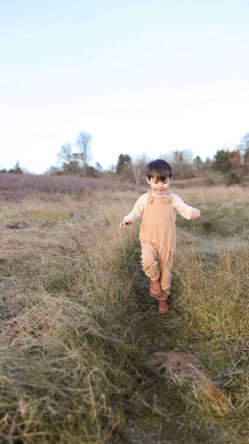 May include: A toddler wearing a light beige long-sleeved shirt underneath tan overalls with knotted shoulder straps and brown leather boots. The clothing is solid-colored and features a simple, functional design suitable for outdoor movement.