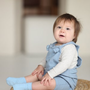 May include: A light blue romper with shoulder ties worn over a long-sleeved cream-colored thermal shirt, paired with matching light blue socks. The outfit features a simple design with a soft texture, providing a comfortable look for a young child.