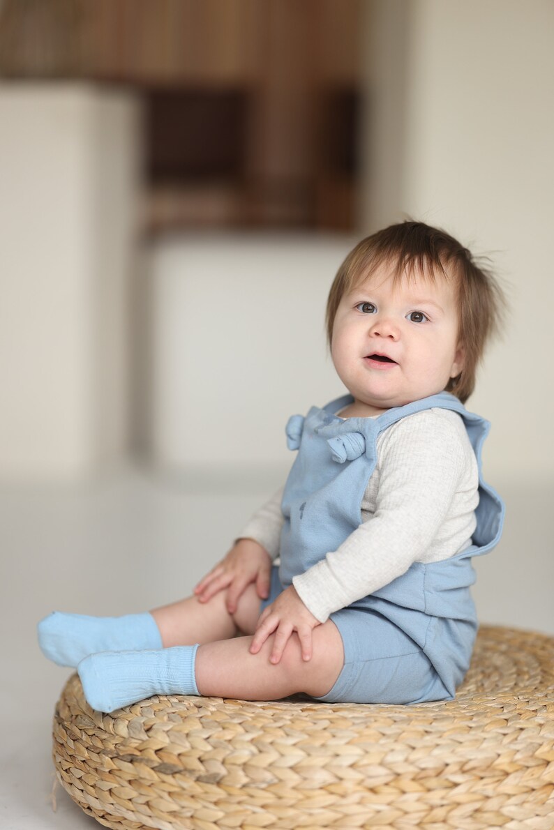 May include: A light blue romper with shoulder ties worn over a long-sleeved cream-colored thermal shirt, paired with matching light blue socks. The outfit features a simple design with a soft texture, providing a comfortable look for a young child.