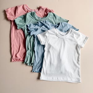 May include: Four ribbed knit t-shirts in pastel colors: pink, green, blue, and white. Each shirt has short sleeves and a round neckline. The shirts are arranged in a diagonal row on a neutral background.
