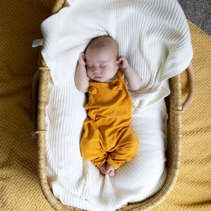 May include: A sleeping infant wearing a mustard yellow overall with knotted shoulder straps, resting inside a woven wicker basket lined with a soft, white, ribbed knit blanket. The basket is placed on a textured mustard yellow fabric surface.