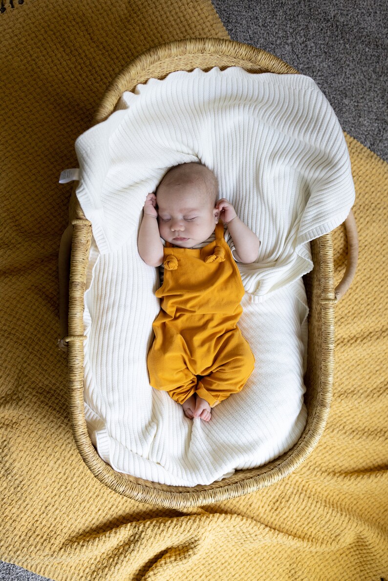 May include: A sleeping infant wearing a mustard yellow overall with knotted shoulder straps, resting inside a woven wicker basket lined with a soft, white, ribbed knit blanket. The basket is placed on a textured mustard yellow fabric surface.
