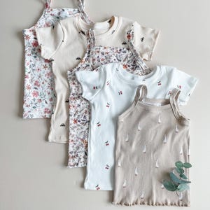 May include: A collection of children's tops in various styles and patterns. The tops include a floral print camisole, a cream-colored short-sleeved shirt with car graphics, a floral print short-sleeved shirt, a white shirt with cherry graphics, and a tan tank top with goose graphics.