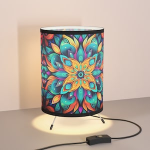 May include: A cylindrical table lamp with a vibrant floral mandala design in shades of orange, teal, and purple. The lamp has a black trim and a three-legged base. The lamp is turned on, casting a warm glow.