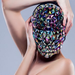 May include: A person wearing a black mask with iridescent triangular sequins covering their face. The mask is held in place by the person's hands.