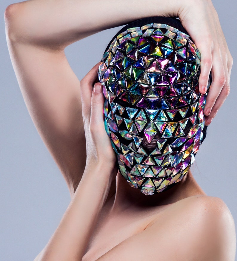 May include: A person wearing a black mask with iridescent triangular sequins covering their face. The mask is held in place by the person's hands.