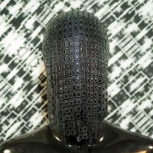 Op de afbeelding: Een zwart gaasmasker met een geometrisch patroon. Het masker is gemaakt van kleine, vierkante metalen stukjes die aan elkaar zijn verbonden. Het masker bedekt het hele hoofd en heeft een lange, stromende franje.