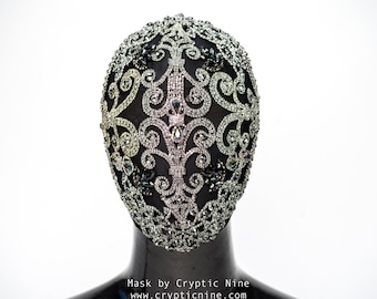 May include: A black masquerade mask with a silver and crystal embellishment design. The mask is on a black mannequin head. The text "Mask by Cryptic Nine www.crypticnine.com" is below the mask.