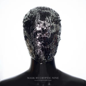 May include: A full face mask covered in black sequins with a silver floral and paisley design. The mask is displayed on a black mannequin head. The text "MASK BY CRYPTIC NINE" and the website address are visible at the bottom.