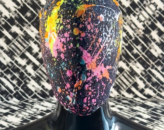 May include: A black face mask with colorful paint splatters in pink, orange, yellow, and green. The mask is made of a stretchy fabric and has a unique design. "MASK BY CRYPTIC NINE www.crypticnine.com" is printed on the bottom of the image.