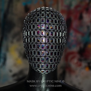 May include: A silver face mask with iridescent squares covering the entire surface. The mask is on a mannequin head. The text "MASK BY CRYPTIC NINE © www.crypticnine.com" is at the bottom of the image.