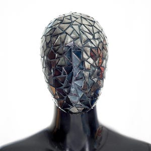 Può includere: Una maschera decorativa con un design a mosaico. La maschera è composta da piccoli pezzi triangolari riflettenti, creando un effetto specchio. La maschera è montata su una testa di manichino nera, su sfondo bianco.