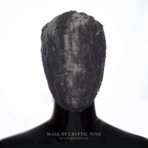 Op de afbeelding: Een zwart masker met een getextureerd oppervlak, dat het hele gezicht bedekt. Het masker wordt getoond op een zwarte mannequinhoofd. De tekst "MASK BY CRYPTIC NINE" en het websiteadres zijn onderaan zichtbaar.