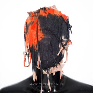 May include: A black and orange mask with shredded fabric details. The mask has a distressed appearance with orange and black dye effects. The bottom of the mask is black and shiny. The text "MASK BY CRYPTIC NINE" is visible at the bottom.