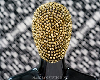 May include: A gold-colored mask with a face-covering design. The mask is covered in small, round, gold-colored studs. The mask is on a black mannequin head. The text "MASK BY CRYPTIC NINE" and "www.crypticnine.com" is printed below the mask.