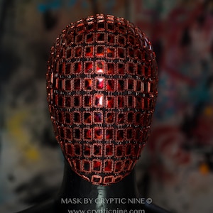 May include: A red face mask with a geometric design made of small, square-shaped pieces. The mask is made of a shiny material and has a reflective surface. The mask is worn on a mannequin's head. The text "MASK BY CRYPTIC NINE © www.crypticnine.com" is printed below the mask.