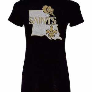 Puede incluir: Camiseta negra con un diseño de strass del estado de Luisiana, la palabra "Saints", una flor de lis y un sombrero de vaquero con la palabra "NOLA" en él.