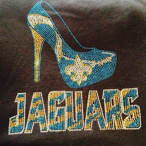 May include: Black fabric featuring a high-heeled shoe design and the word "JAGUARS", all embellished with teal, gold, and white beads. The shoe design includes a fleur-de-lis and a curved strap.