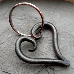 Pode incluir: Um chaveiro de metal em forma de coração forjado à mão, com um anel de chave prateado. O coração é de metal escuro e tem um design em espiral na parte superior.