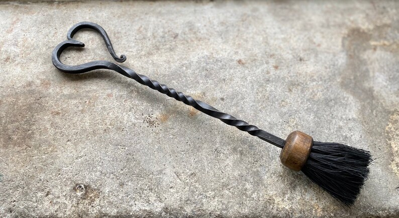 Pode incluir: Uma escova de lareira de metal preto com uma pega torcida e uma parte superior em forma de cora&ccedil;&atilde;o. A escova tem uma pega de madeira e cerdas pretas.