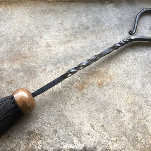 Pode incluir: Uma escova de lareira de metal preto com uma al&ccedil;a torcida e uma parte superior em forma de cora&ccedil;&atilde;o. A escova tem uma al&ccedil;a de madeira e cerdas pretas.