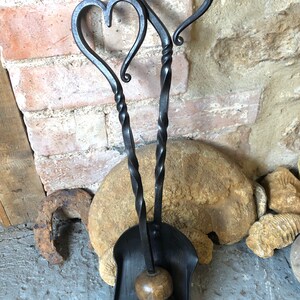 Pode incluir: Um conjunto de ferramentas para lareira de metal preto com al&ccedil;as em forma de cora&ccedil;&atilde;o. O conjunto inclui uma p&aacute;, uma escova e um atizador. As ferramentas s&atilde;o torcidas e t&ecirc;m um aspeto r&uacute;stico.