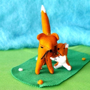 May include: Two orange felt fox figures, one large and one small, stand on a green felt base. The larger fox has a white-tipped tail and black whiskers. The background is a light blue and green felt landscape.