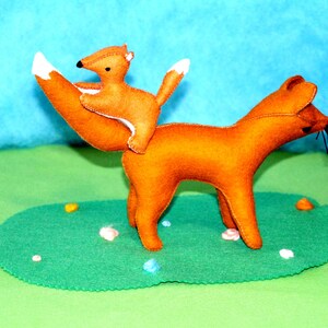 May include: A handmade felt fox toy with a smaller fox riding on its back. The foxes are orange with white-tipped tails and black details. They are positioned on a green felt base with colorful accents, set against a blue backdrop.