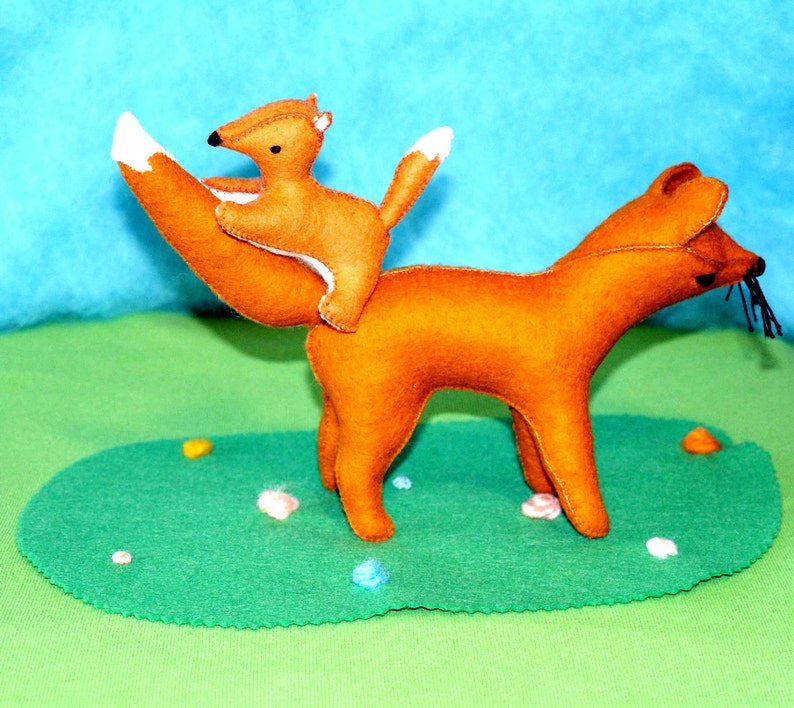 May include: A handmade felt fox toy with a smaller fox riding on its back. The foxes are orange with white-tipped tails and black details. They are positioned on a green felt base with colorful accents, set against a blue backdrop.