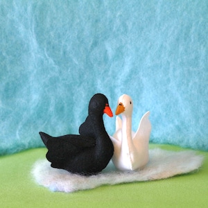 May include: A black swan and a white swan facing each other on a white fluffy surface. The swans are made of felt and have orange beaks.