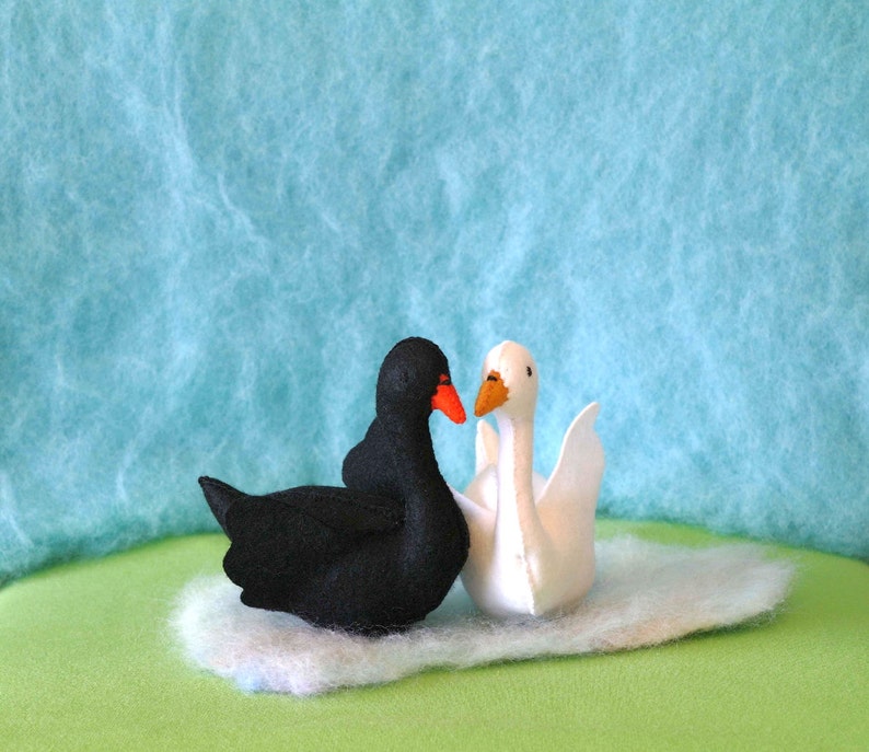 May include: A black swan and a white swan facing each other on a white fluffy surface. The swans are made of felt and have orange beaks.