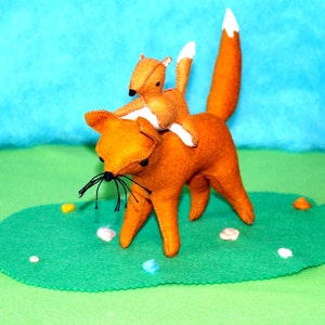 May include: A handmade felt fox carrying a smaller felt fox on its back. The foxes are orange with white-tipped tails and black whiskers. They are set on a green felt base with a blue background.