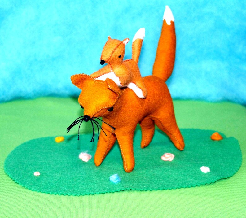 May include: A handmade felt fox carrying a smaller felt fox on its back. The foxes are orange with white-tipped tails and black whiskers. They are set on a green felt base with a blue background.