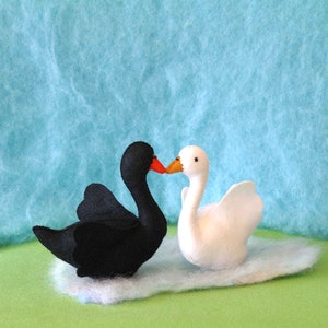 May include: A black swan and a white swan with orange beaks are kissing. The swans are made of felt and are sitting on a white fluffy surface.