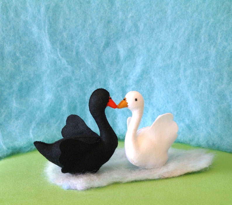 May include: A black swan and a white swan with orange beaks are kissing. The swans are made of felt and are sitting on a white fluffy surface.