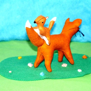 May include: Handmade felt fox toy with a smaller fox riding on its back. The foxes are orange with white accents on their tails and ears. They are positioned on a green felt base with decorative elements, against a blue and green background.