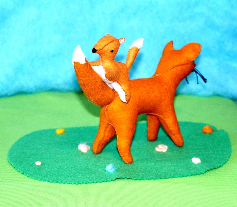 May include: Handmade felt fox toy with a smaller fox riding on its back. The foxes are orange with white accents on their tails and ears. They are positioned on a green felt base with decorative elements, against a blue and green background.