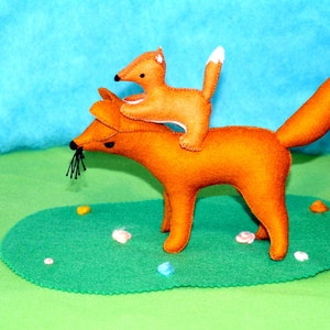 May include: A felt fox toy with a smaller fox riding on its back. The foxes are orange with white-tipped tails and black whiskers. They are set against a green and blue backdrop, creating a whimsical scene.